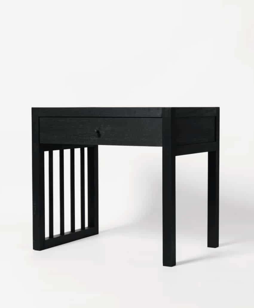 A minimalist black wooden desk with four legs, a single drawer, and vertical slats on the left side, set against a plain white background.