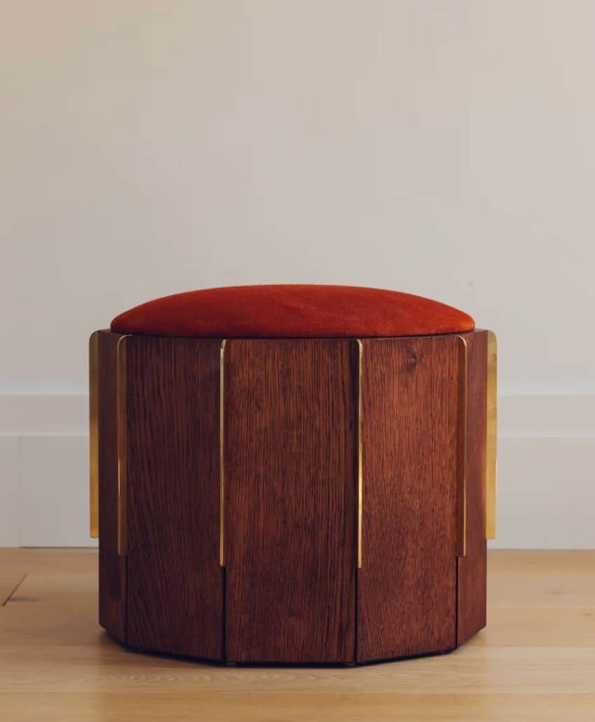 A wooden stool with vertical gold accents and an orange-cushioned seat sits on a light wooden floor against a plain, light-colored wall.