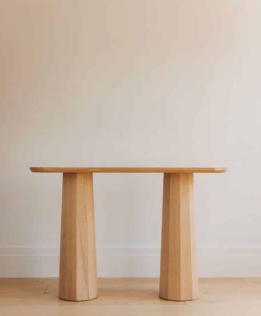 A minimalist wooden table with a flat rectangular top and two wide, tapered cylindrical legs, placed against a plain off-white wall on a light wood floor.