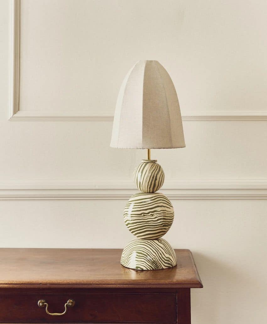 A table lamp with a beige, fabric lampshade and a stacked, round ceramic base featuring a black and white zebra stripe pattern sits on a polished wooden table against a cream-colored wall.