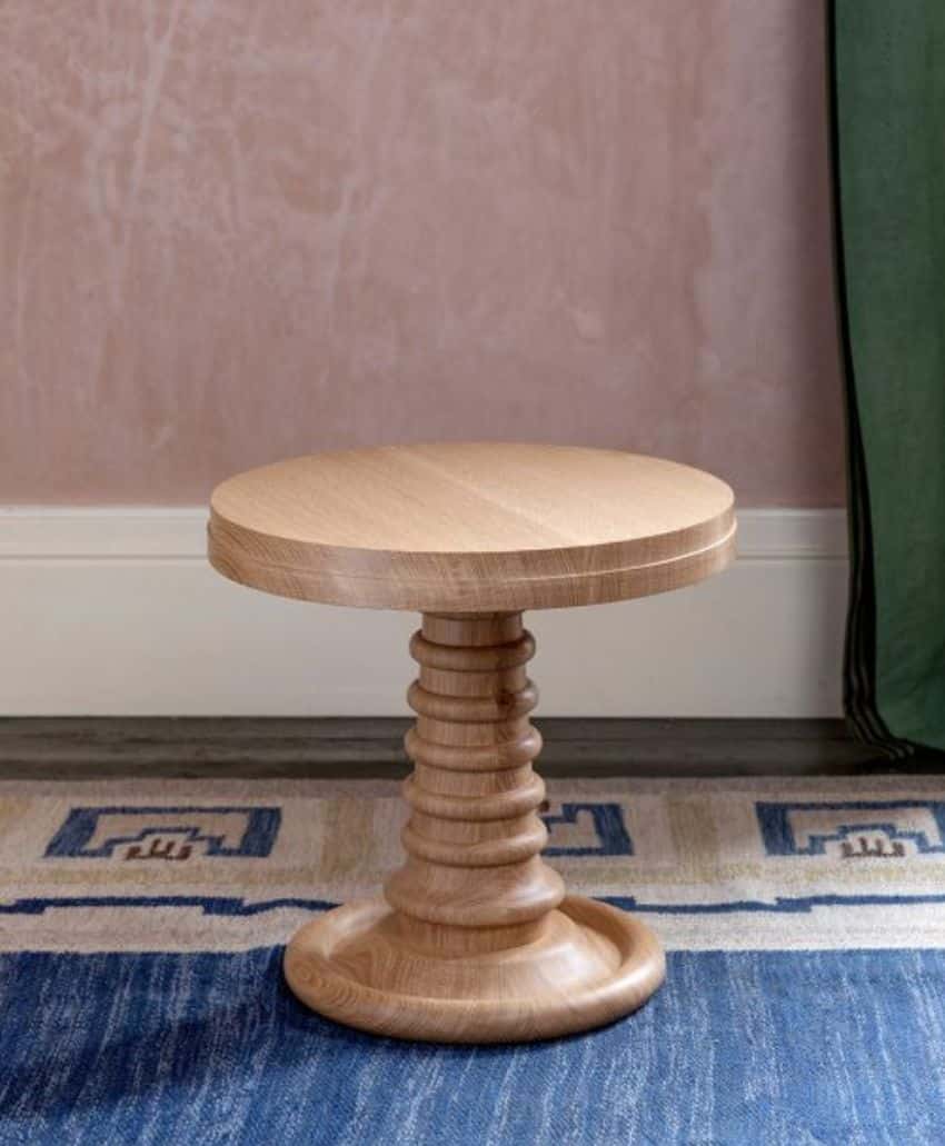 A small, round wooden side table with a sculpted, ridged pedestal base sits on a geometric blue and beige rug against a pale pink wall. A green curtain is partially visible to the right.