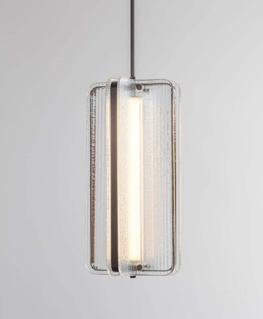 A modern pendant light with a vertical tubular bulb, enclosed by two rectangular, textured glass panels with rounded edges, hung from a black cord against a plain light background.