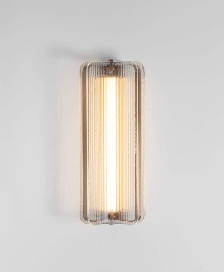 A modern wall sconce with a vertical, rectangular frosted glass cover and a warm light glowing from within, mounted on a plain white wall.