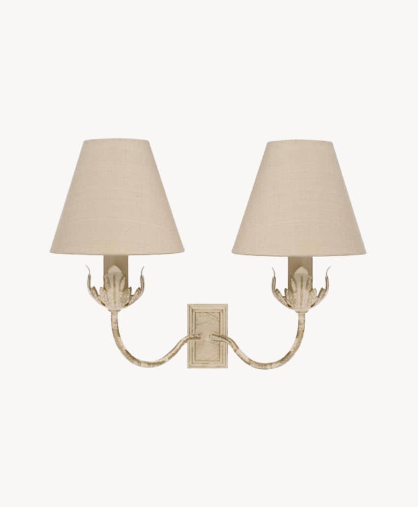 A wall-mounted, two-arm light fixture with beige fabric lampshades and a decorative, textured metal base in a cream color.