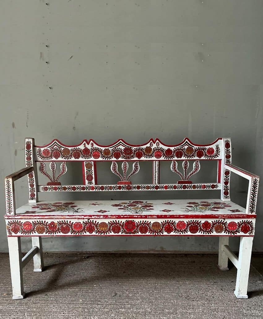 A white wooden bench with red and yellow floral patterns and decorative motifs painted on the backrest, seat, and arms, set against a plain gray wall.