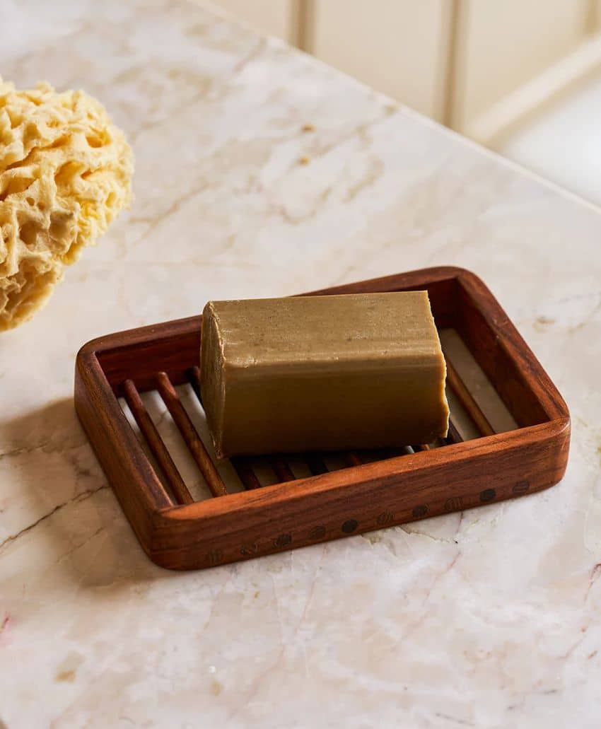 A rectangular bar of brown soap sits on a wooden soap dish with slats, placed on a marble countertop. A natural yellow sponge is partially visible in the background.