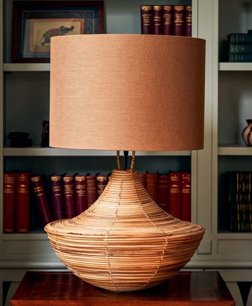 A woven rattan table lamp with a wide, round base and a large beige lampshade sits on a wooden table in front of bookshelves filled with red and blue books.