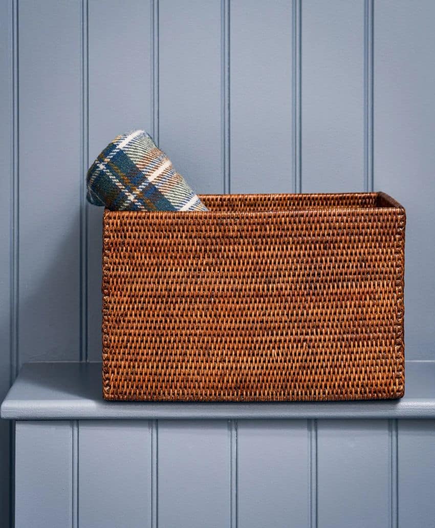 A rectangular wicker basket sits on a blue shelf against a blue paneled wall, with a rolled-up plaid fabric partly visible inside the basket.