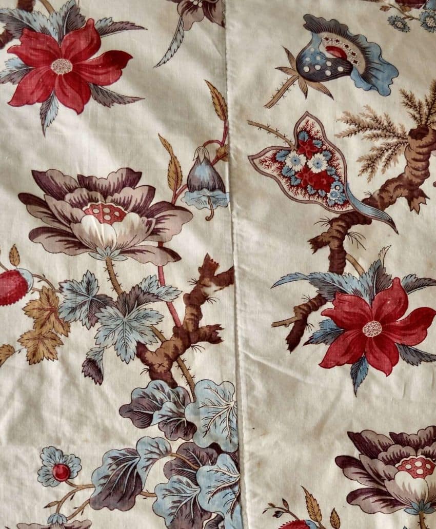 A fabric with a floral and bird print in shades of red, blue, brown, and beige, featuring large flowers, leaves, and stylized birds against a light background with a visible central seam.