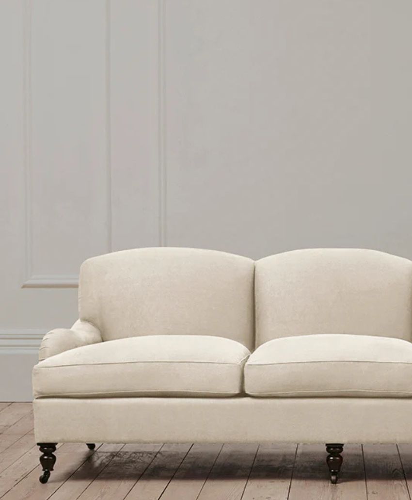 A cream-colored upholstered loveseat with dark wooden legs on a light wooden floor, positioned against a plain, light gray wall with decorative molding.