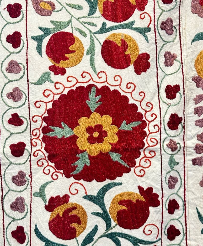 Embroidered textile with red, yellow, and green floral and pomegranate motifs, featuring a large central flower surrounded by decorative borders on a white fabric background.