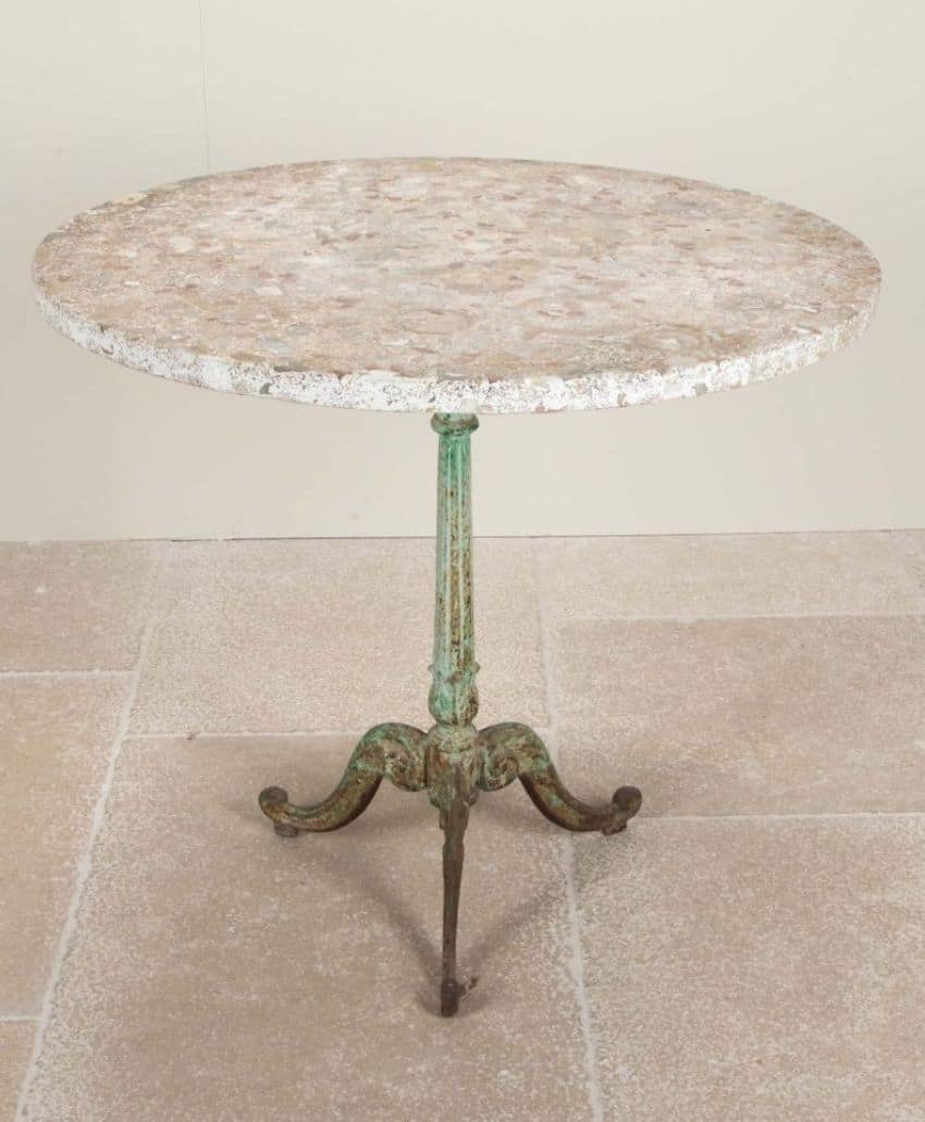 A round stone-top table with a weathered surface, supported by a slender, ornate green metal pedestal and three curved legs, standing on a tiled floor against a plain wall.