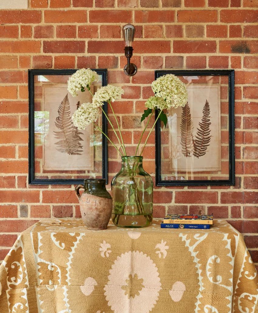 A table with a patterned beige and white cloth holds a green glass vase of white flowers and a rustic jug. Behind, a brick wall displays two framed fern prints and an exposed bulb sconce above.