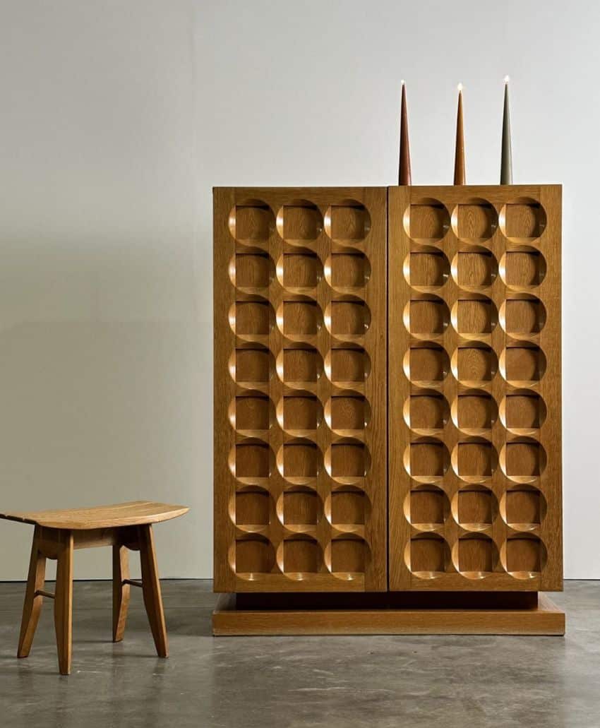 A wooden cabinet with a geometric, rounded square pattern on its doors stands next to a small wooden stool with angled legs. Three tall, cone-shaped objects sit on top of the cabinet. The setting has a concrete floor and plain wall.