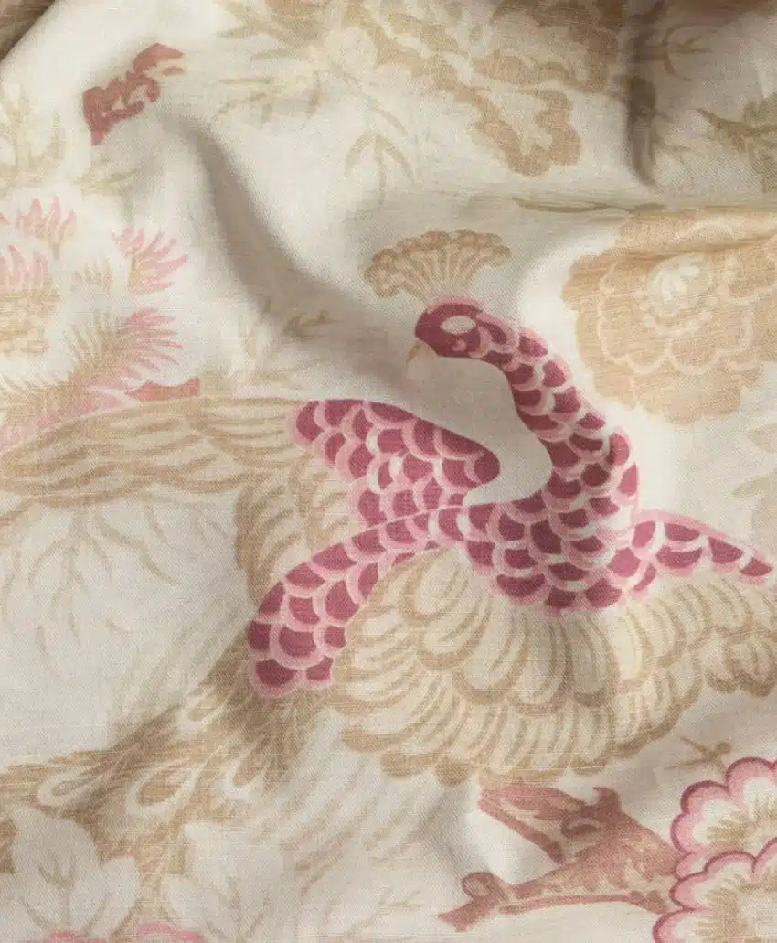 A close-up of fabric with a decorative pattern featuring a pink and beige peacock surrounded by floral designs on a light background.