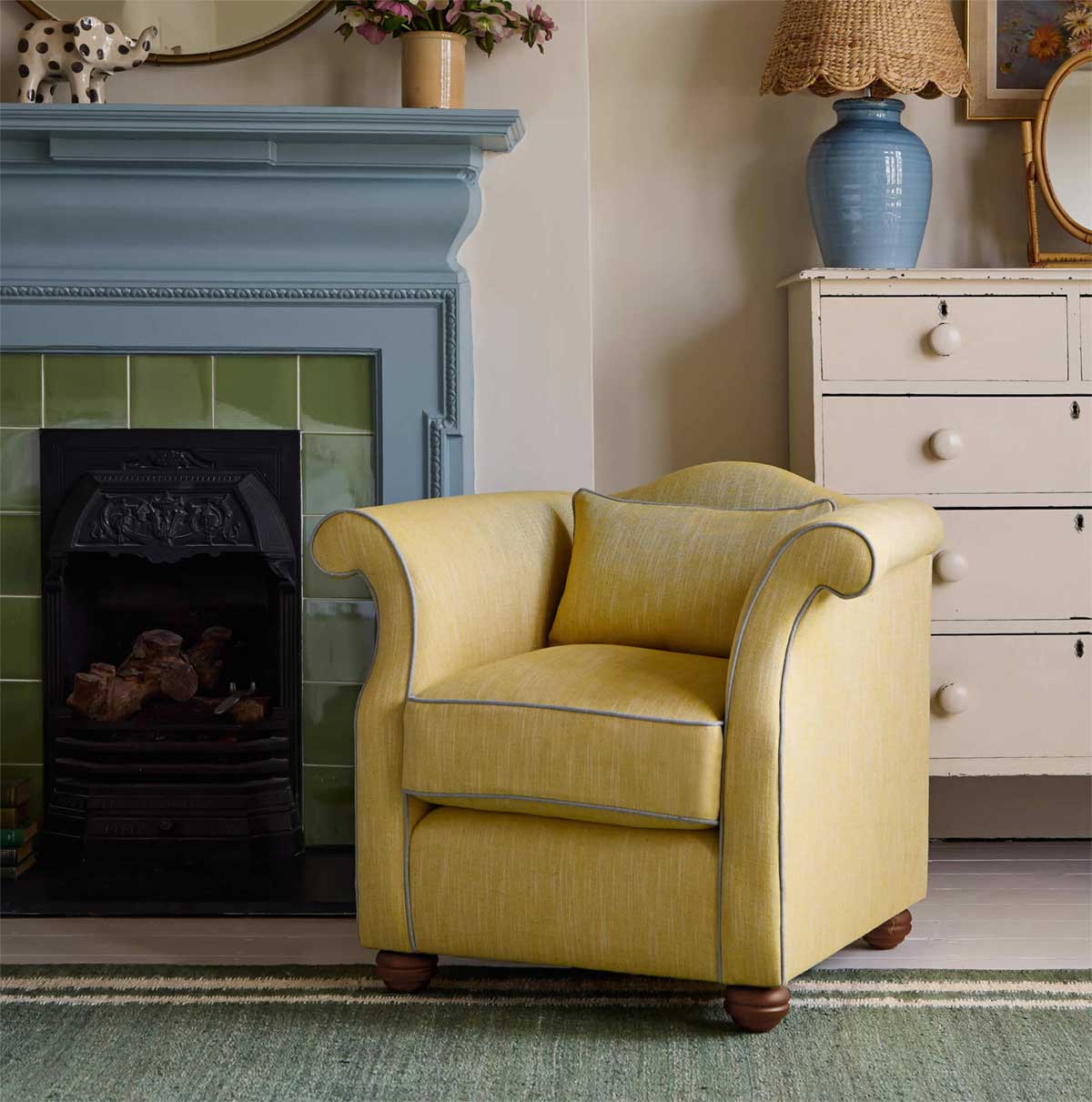 A cozy yellow armchair with rolled arms sits on a green rug beside a blue fireplace and a cream dresser, with vases and a lamp on top, in a warmly decorated room.