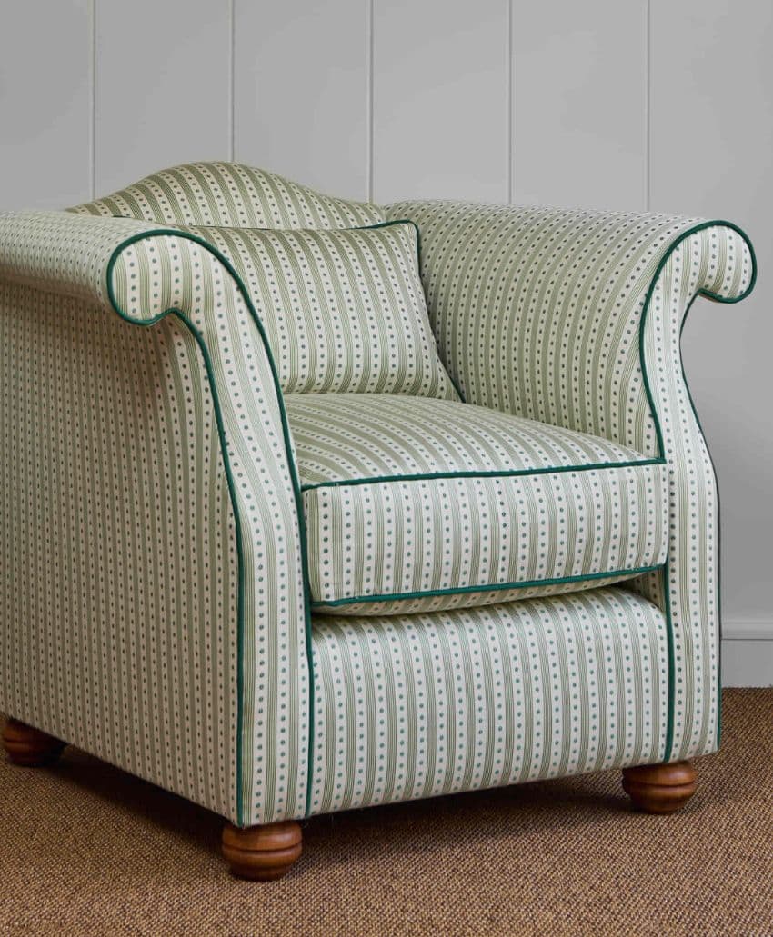 A green and white striped armchair with rolled arms, a matching cushion, dark green piping, and wooden legs, placed on a brown carpet in front of a white paneled wall.