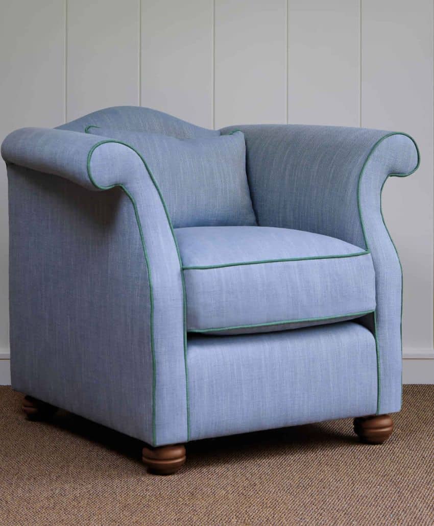 A light blue upholstered armchair with rounded arms and dark wooden feet, featuring green piping along the edges, sits on a tan carpet against a white-paneled wall.