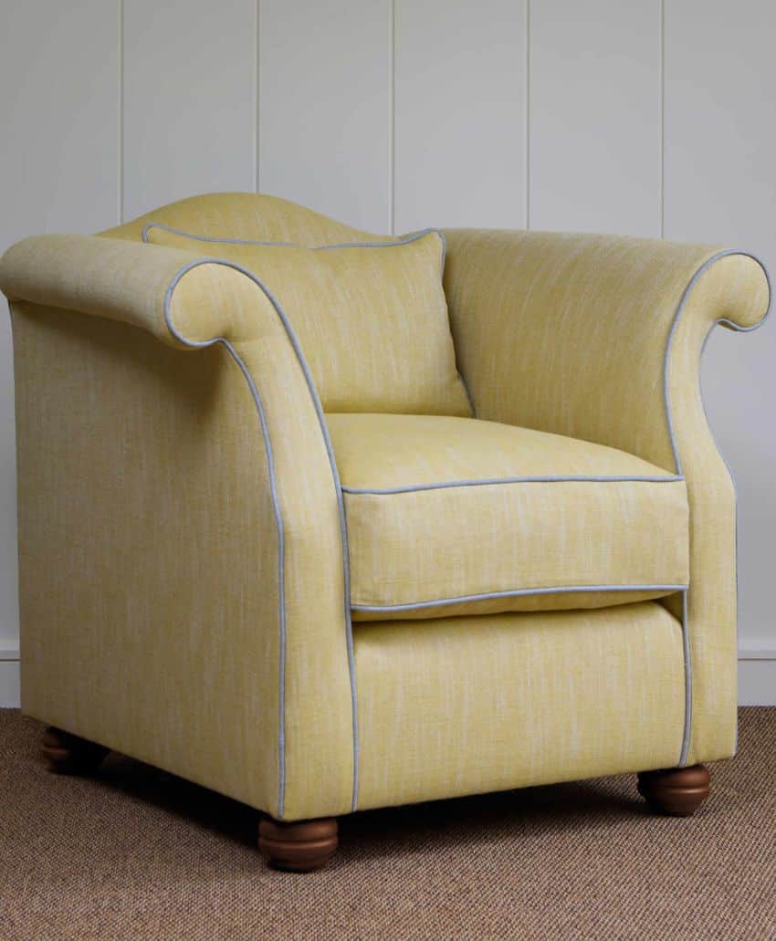 A yellow upholstered armchair with rolled arms, light blue piping, a cushioned seat, and wooden feet, placed on a brown carpet in front of a white paneled wall.