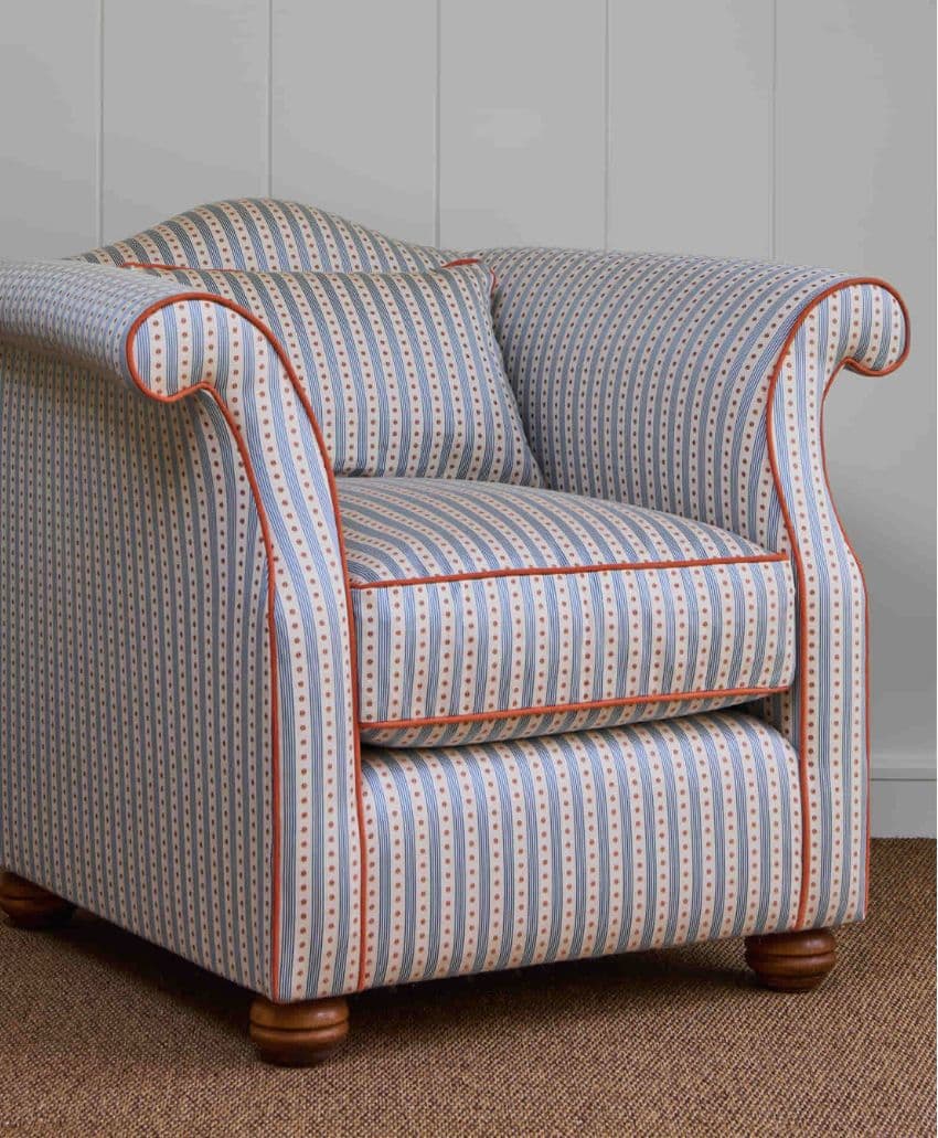 A cushioned armchair with rolled arms, upholstered in blue-and-white striped fabric with small red and beige patterns, and red piping. The chair sits on short wooden legs on a brown rug against a light paneled wall.