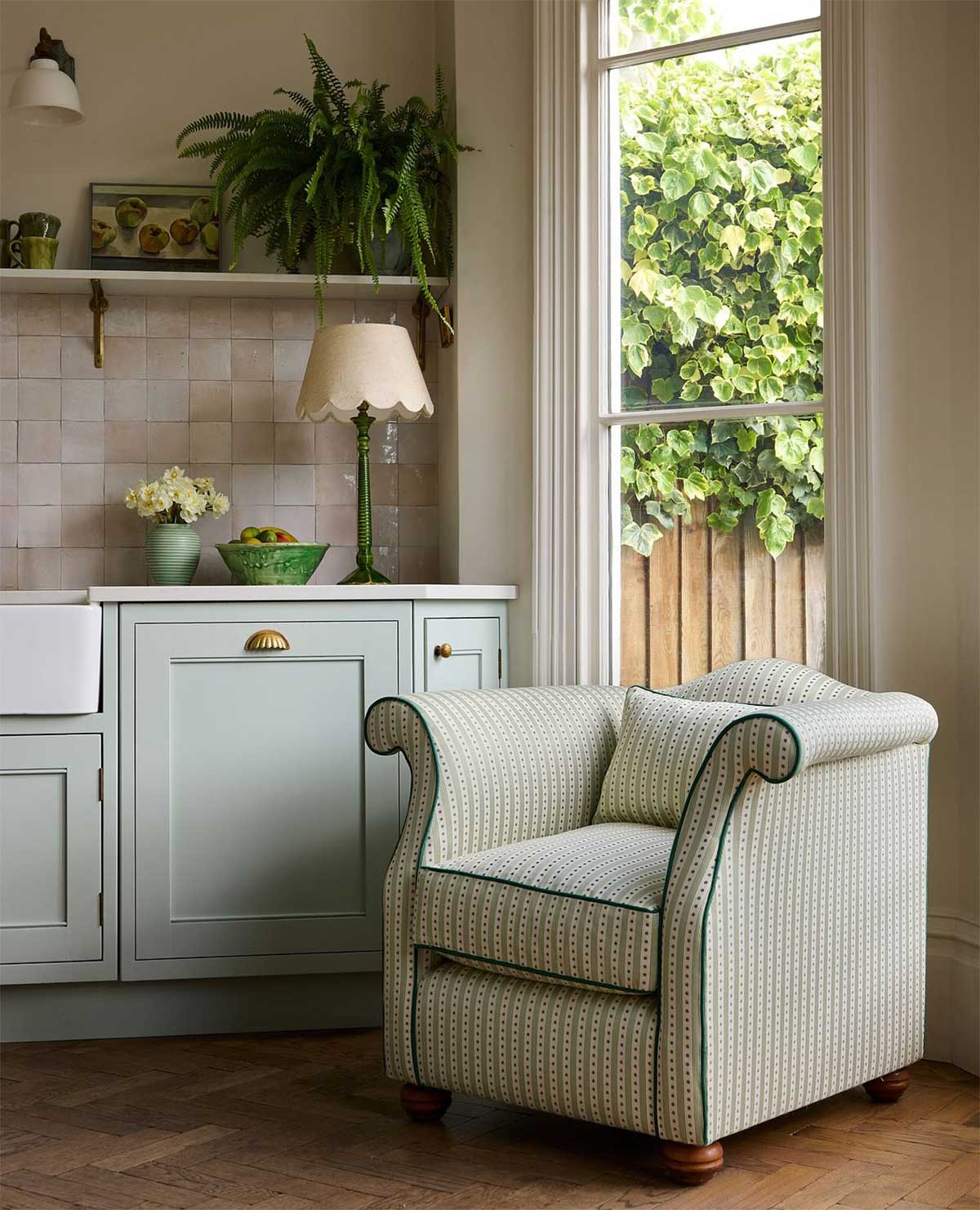 A light green patterned armchair sits in a cozy kitchen corner near a window, with pale green cabinets, a lamp, and potted plants on the counter, and leafy greenery visible outside.