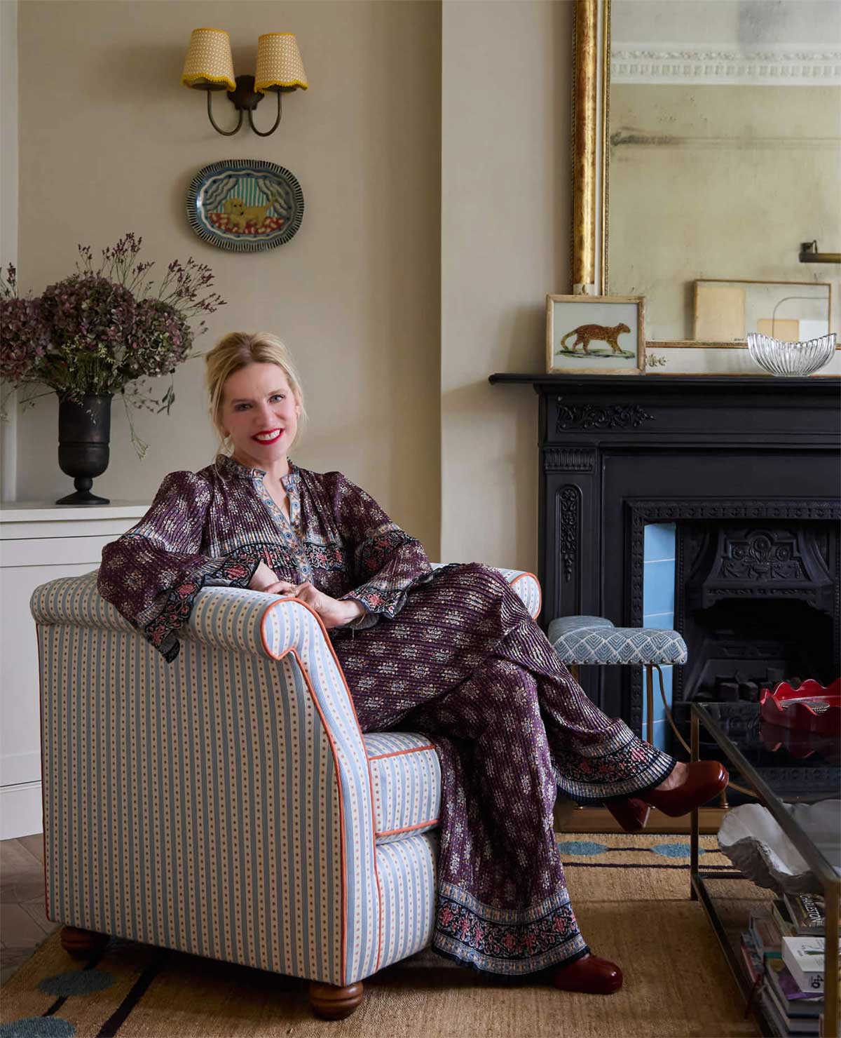 A woman with blonde hair, wearing a patterned outfit, sits relaxed in a striped armchair in a cozy living room with a fireplace, a large mirror, and decorative wall art.