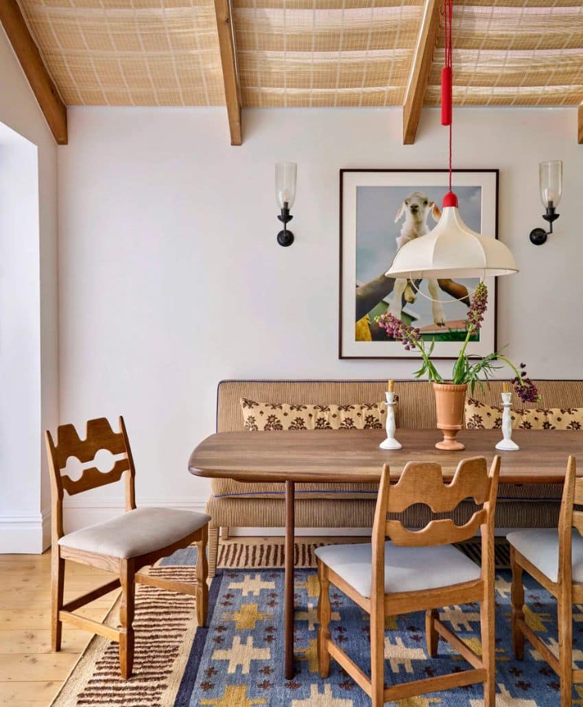 A cozy dining area features a wooden table, three mid-century chairs, a patterned rug, a cushioned bench, a framed goat photo on the wall, and a pendant light hanging from a beamed ceiling. Candles and flowers decorate the table.