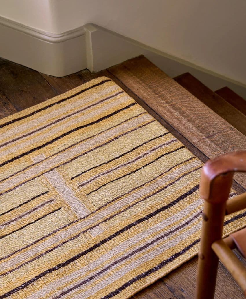 A woven yellow and brown striped rug lies at the top of wooden stairs next to a wooden chair in a cozy interior setting.