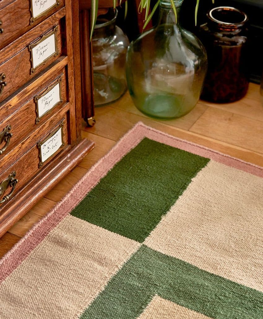 A close-up of a patterned rug with green and beige blocks and a mauve border on a wooden floor, next to a vintage wooden cabinet and two large glass jars.