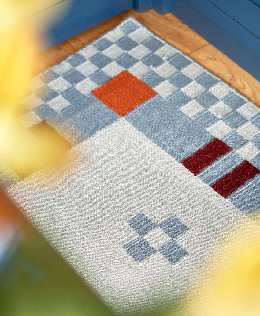 A close-up of a geometric-patterned rug with blue, white, gray, red, and orange squares and rectangles, placed on a wooden floor beside blue furniture. Blurred yellow objects appear in the foreground.