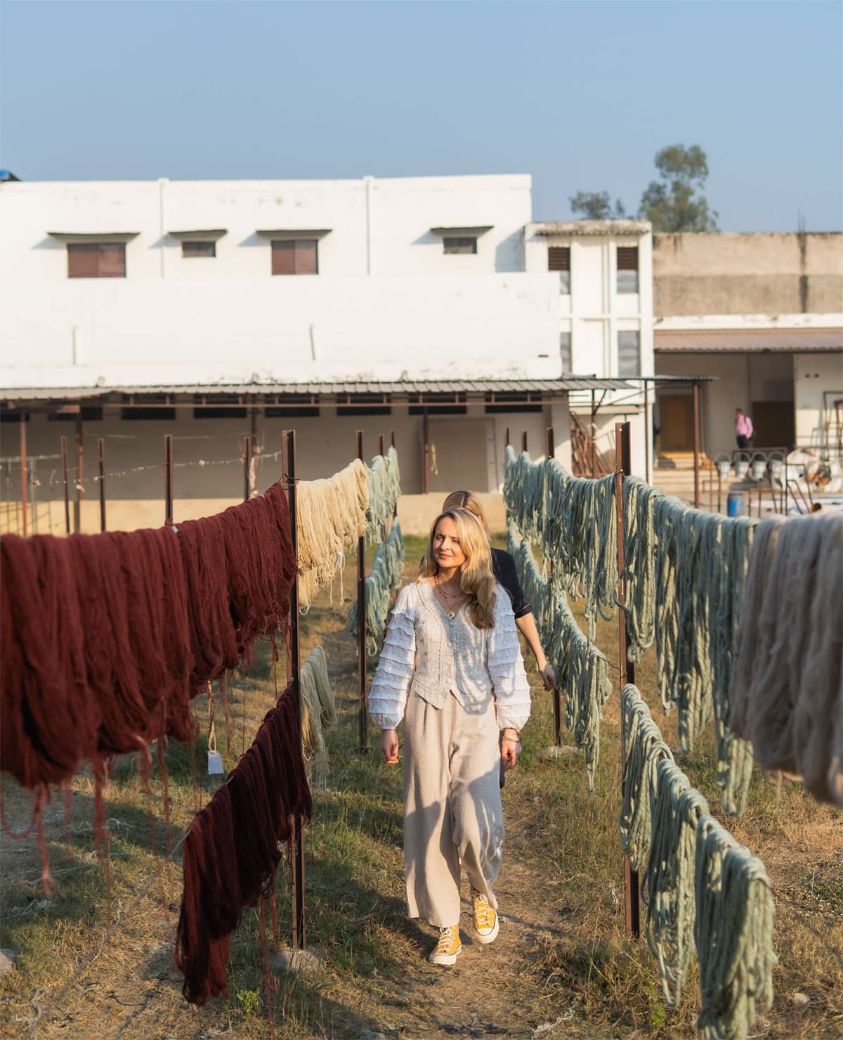 A woman walks between rows of colored yarn hanging to dry outdoors, with white and brown buildings in the background under a clear blue sky.