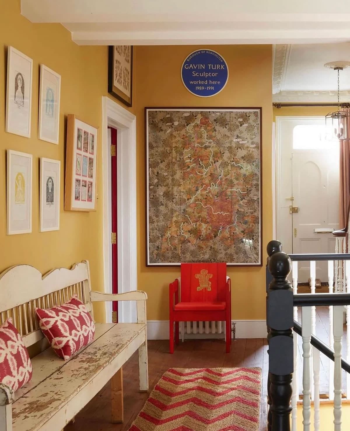 A warm yellow hallway with a rustic bench, red-patterned pillows, framed art on the wall, a large map, a small red chair, and a chevron rug. Sunlight shines through a door in the background.