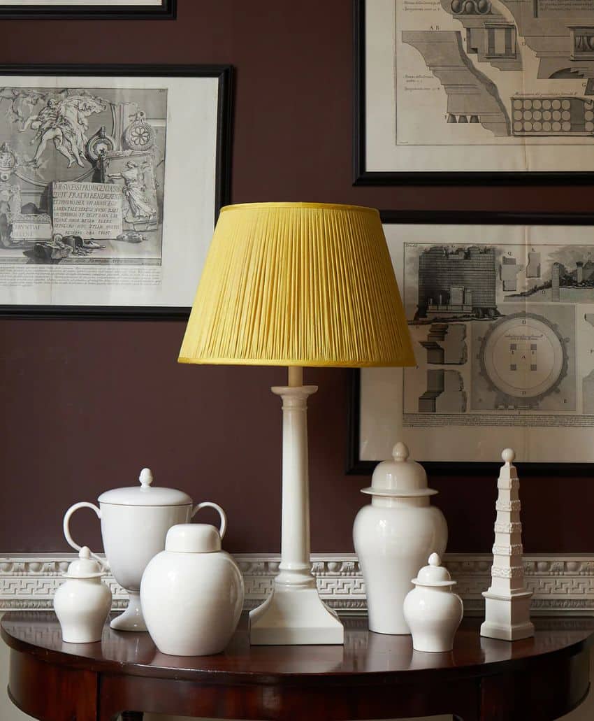 A white table lamp with a yellow pleated shade sits on a wooden table, surrounded by white ceramic jars and decorative objects. Framed architectural prints hang on the dark brown wall behind.