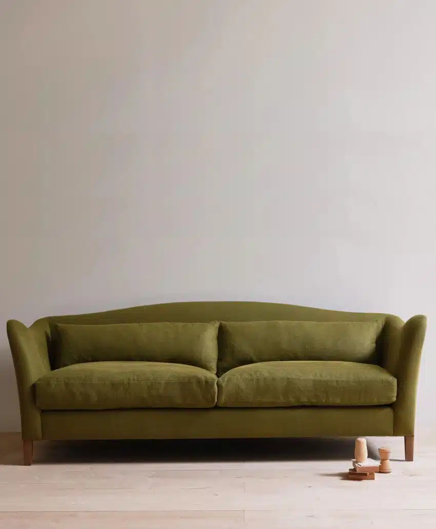 A green velvet sofa with two seat cushions is placed against a plain, light-colored wall on a wooden floor. Three small wooden toys are arranged on the floor near the bottom right corner of the sofa.