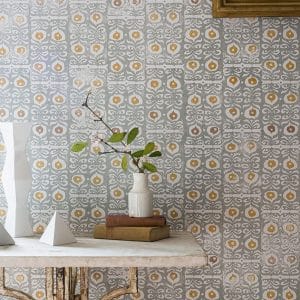 A patterned wall with gray and yellow tile design, a rustic table with a white vase holding leafy branches, two white geometric sculptures, and stacked books. Part of a framed picture is visible in the top right corner.