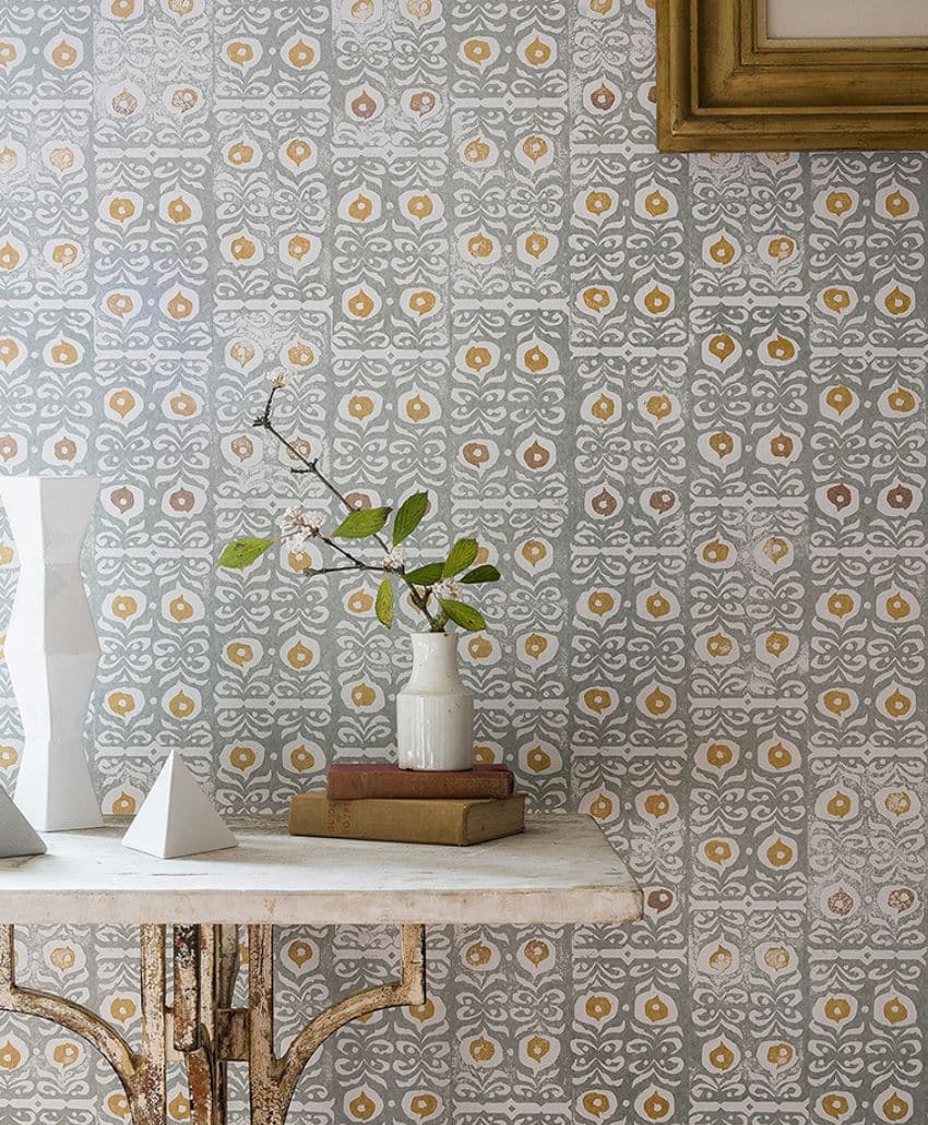 A patterned wall with gray and yellow tile design, a rustic table with a white vase holding leafy branches, two white geometric sculptures, and stacked books. Part of a framed picture is visible in the top right corner.