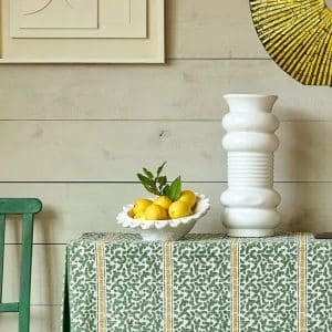 A decorative table with a green patterned cloth holds a white vase and a white bowl filled with yellow lemons and leaves. A green chair is beside the table; art and a round yellow mirror hang on a light wood wall.