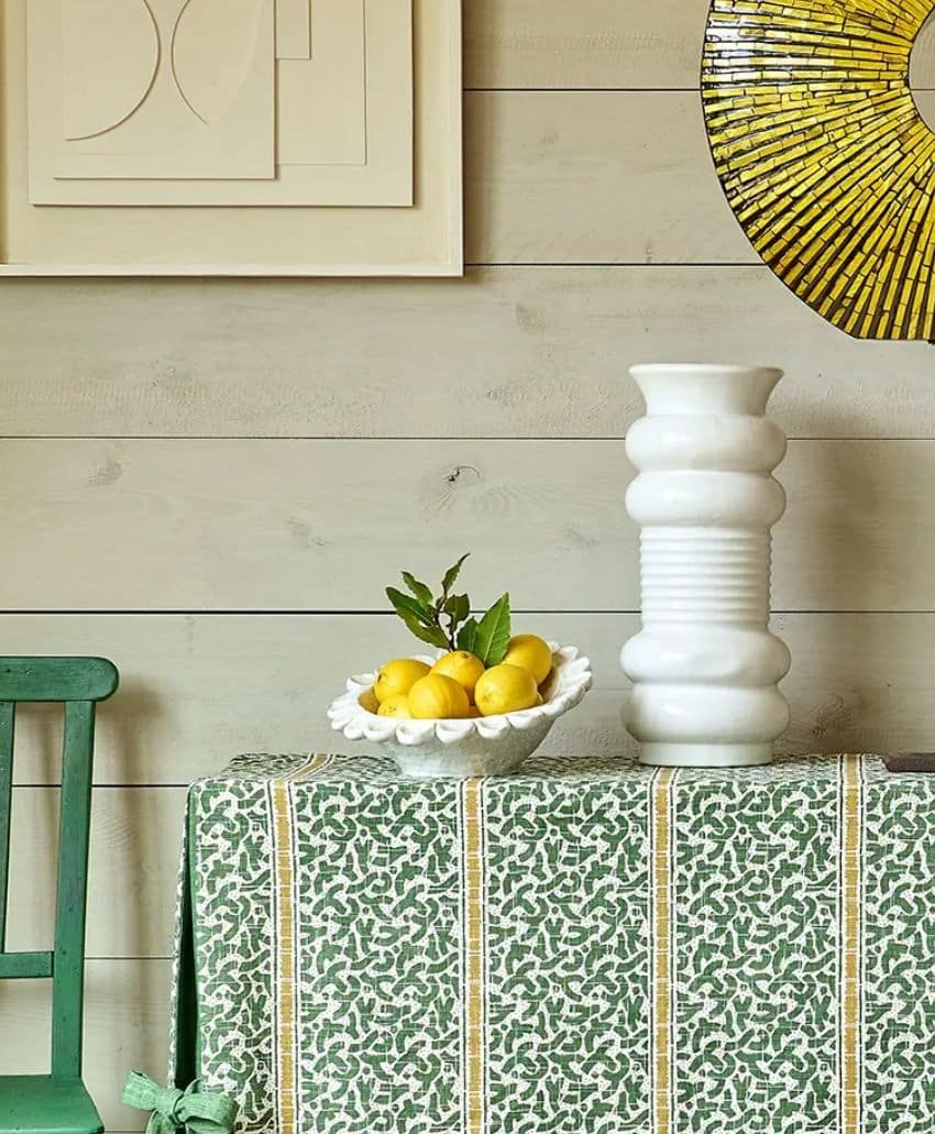 A decorative table with a green patterned cloth holds a white vase and a white bowl filled with yellow lemons and leaves. A green chair is beside the table; art and a round yellow mirror hang on a light wood wall.