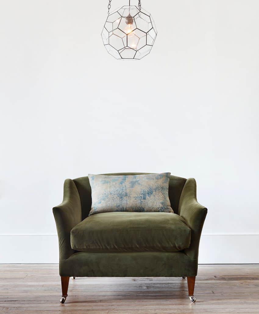 A green velvet armchair with wooden legs sits on a wooden floor against a white wall. A light blue patterned cushion rests on the chair, and a geometric pendant light hangs above.
