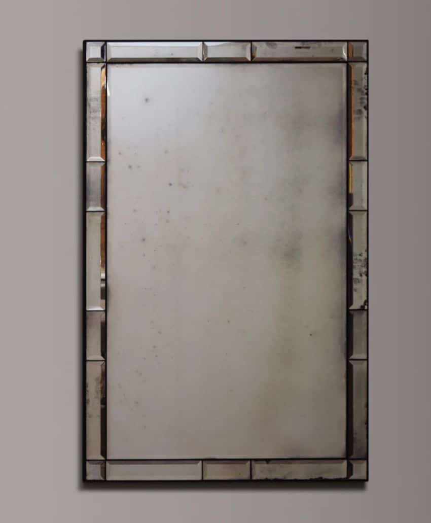 A rectangular, antique-style mirror with a wide, beveled metallic frame set against a plain gray wall. The glass has a slightly foggy or distressed appearance, adding a vintage touch.