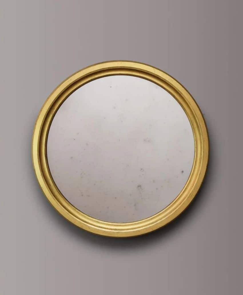 A round mirror with a gold frame is mounted on a plain, light gray wall. The mirror’s surface appears slightly cloudy with small spots, suggesting age or wear.