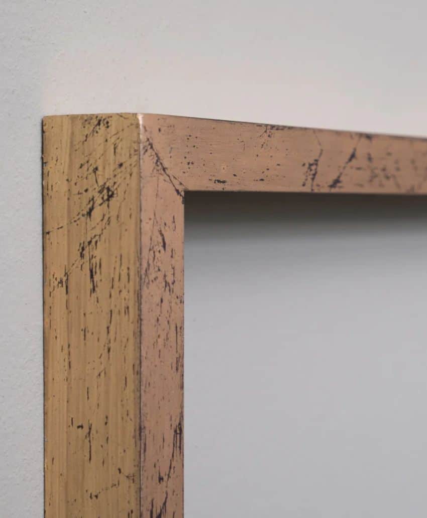 Close-up of the corner of a wooden or metal picture frame with a distressed, gold finish, showing visible black scratch marks and texture against a plain, light-colored wall.