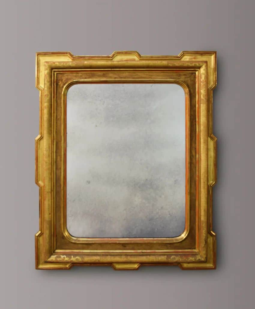 A rectangular antique mirror with a wide, ornate gold frame featuring beveled edges and decorative corners, mounted on a plain gray wall.