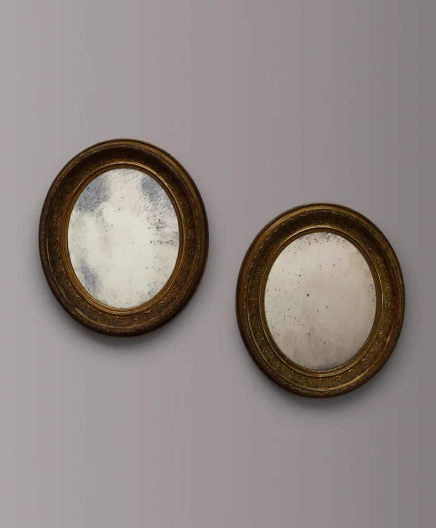 Two round, antique mirrors with ornate, dark gold frames hang on a plain gray wall. The mirrors appear slightly aged and tarnished, showing signs of wear and spots on their surfaces.