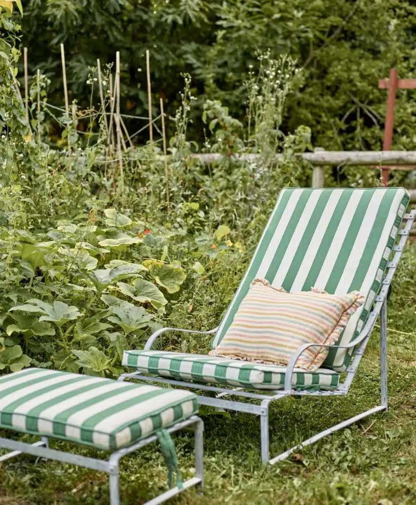 A green and white striped outdoor lounge chair with a matching footrest and a striped pillow sits on grass in a lush garden surrounded by leafy plants and wooden garden supports.