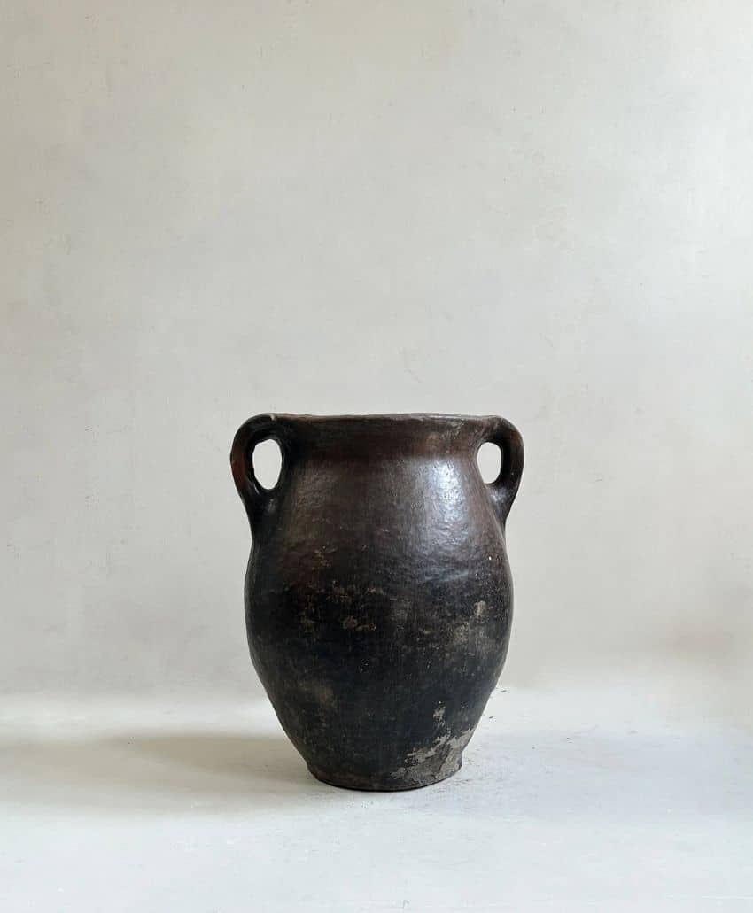 A large, dark, weathered clay pot with two rounded handles sits on a plain, light-colored surface against a neutral background.