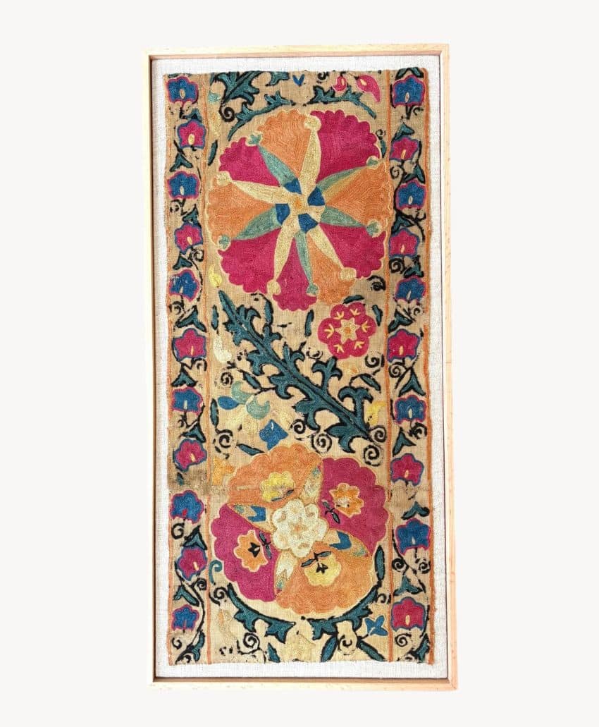 A rectangular embroidered textile with intricate floral patterns in red, yellow, orange, and teal, bordered with decorative motifs, framed in a light wooden frame against a neutral background.