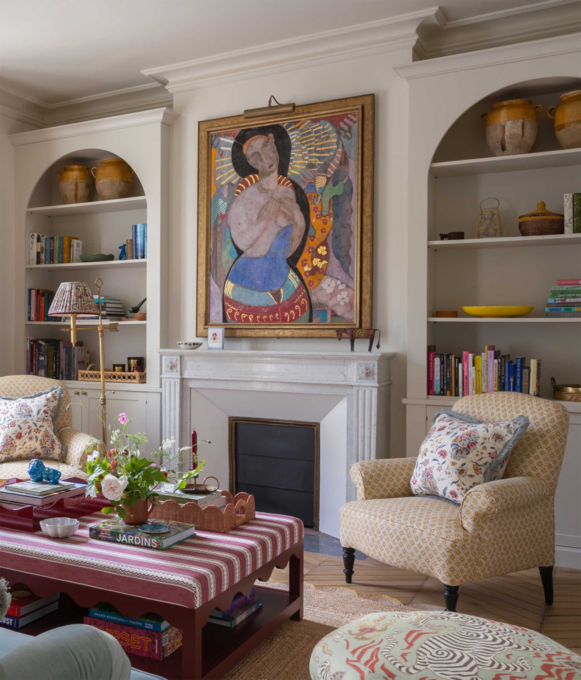 A cozy living room with built-in shelves, a white fireplace, patterned armchairs, a striped coffee table, and a colorful abstract portrait hanging above the mantel. Books, pottery, and flowers add decorative touches.