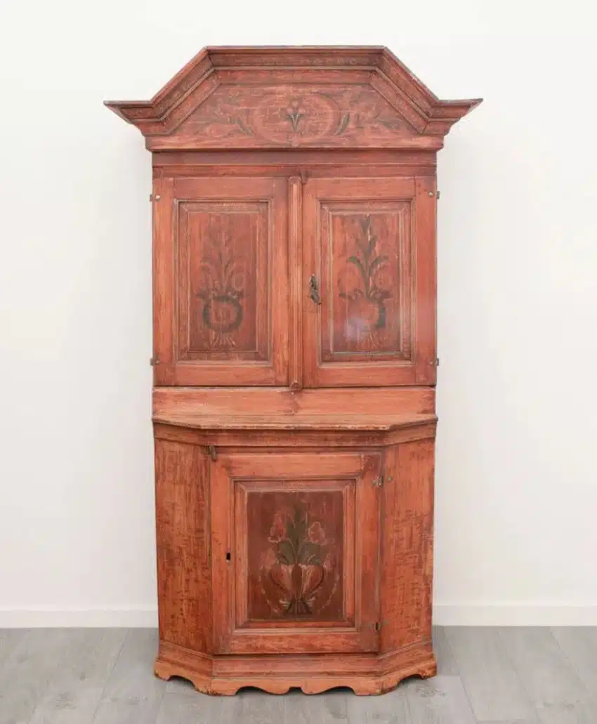 Antique wooden cabinet with a reddish-brown finish, decorative floral carvings on the doors, and an angled top. The cabinet has upper and lower compartments and stands against a white wall on a gray floor.