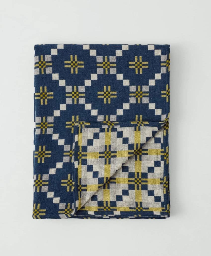 A folded blanket with a geometric pattern in navy blue, white, and yellow, featuring squares and cross shapes, lying on a plain white background. One corner is flipped to reveal the backside of the pattern.